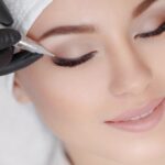 A relaxed woman lying on couch at beauty salon with closed eyes while a professional wearing black gloves is making permanent eyeliner make up.