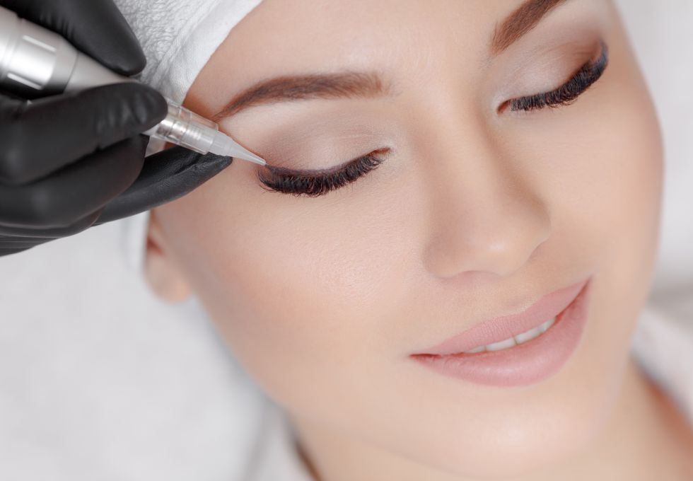 A relaxed woman lying on couch at beauty salon with closed eyes while a professional wearing black gloves is making permanent eyeliner make up.